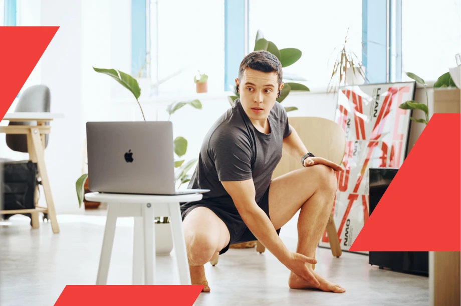 Man in athletic wear following an online workout on a laptop while stretching indoors in a bright, plant-filled room.
