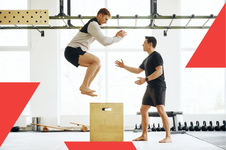 A man is performing a box jump exercise in a gym while another man, possibly a trainer, stands nearby with arms extended to assist or guide him. The gym has large windows, fitness equipment like kettlebells in the background, and both men are barefoot and wearing athletic clothing.