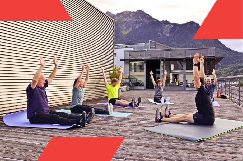 Group of adults performing seated stretching exercises on yoga mats on a rooftop deck with mountains in the background.