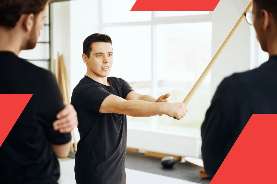 A fitness instructor is demonstrating an exercise using a wooden stick to two participants in a gym setting. He holds the stick with both hands, showing proper technique, while the others attentively watch. The gym is brightly lit with large windows and training equipment in the background.