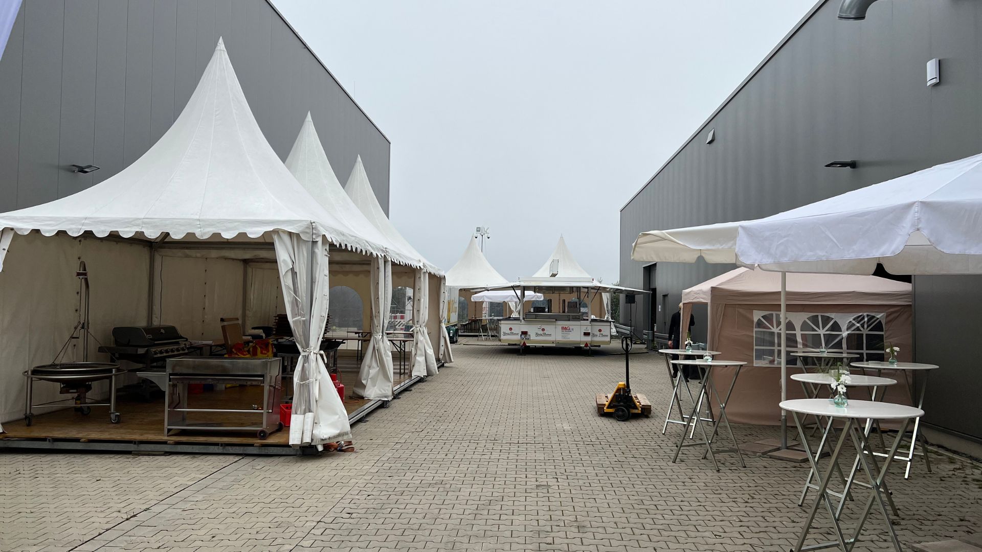 Aufgebautes Außenevent mit weißen Pagodenzelten, Stehtischen und Grillstation zwischen zwei Hallen