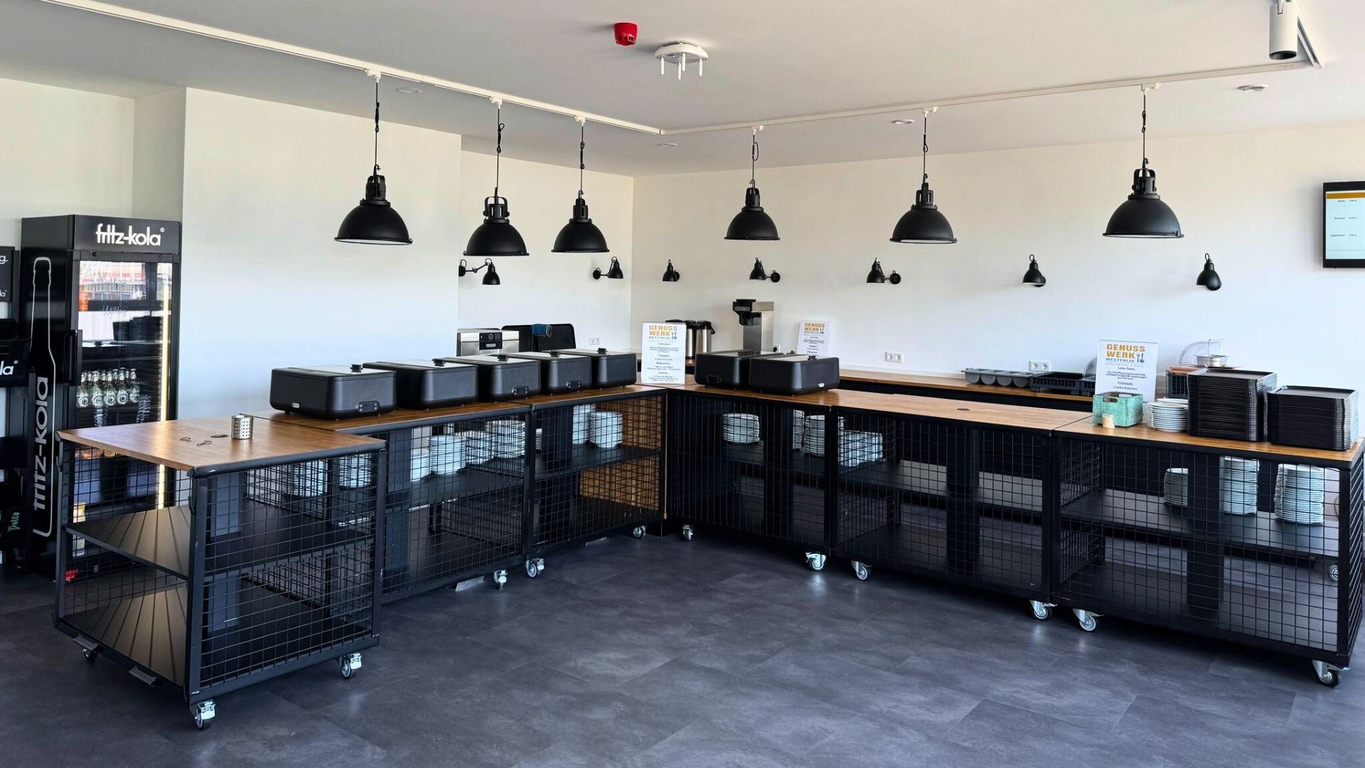 Modernes, leeres Buffet mit schwarzen Metallregalen, Holzplatten, Wärmestation und Getränkekühlschrank in heller Kantine