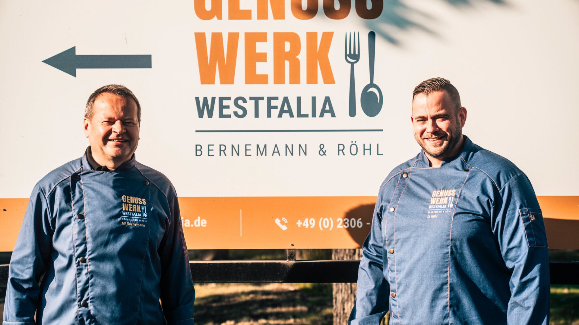 Geschäftsführer in blauen Jacken stehen lächelnd vor einem großen Genusswerk-Westfalia-Schild