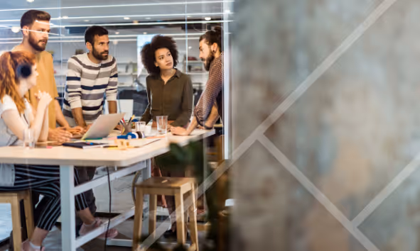 A team of people in a work environment standing around a table having a discussion - First Technology Cyber Security Solutions