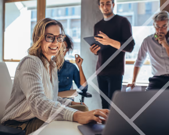 A smiling woman in the foreground working on a laptop with other work colleages in the background