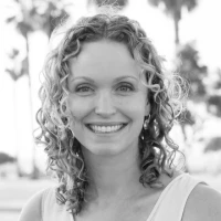 A black and white photo of a woman with curly hair, smiling, wearing a sleeveless top, with trees in the blurred background.