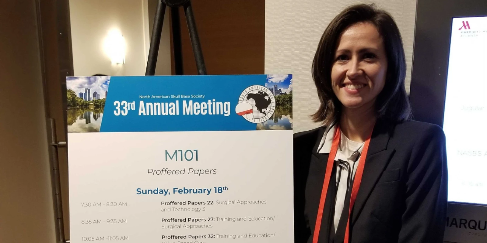 A woman standing next to a sign for the 33rd Annual Meeting of the North American Skull Base Society.