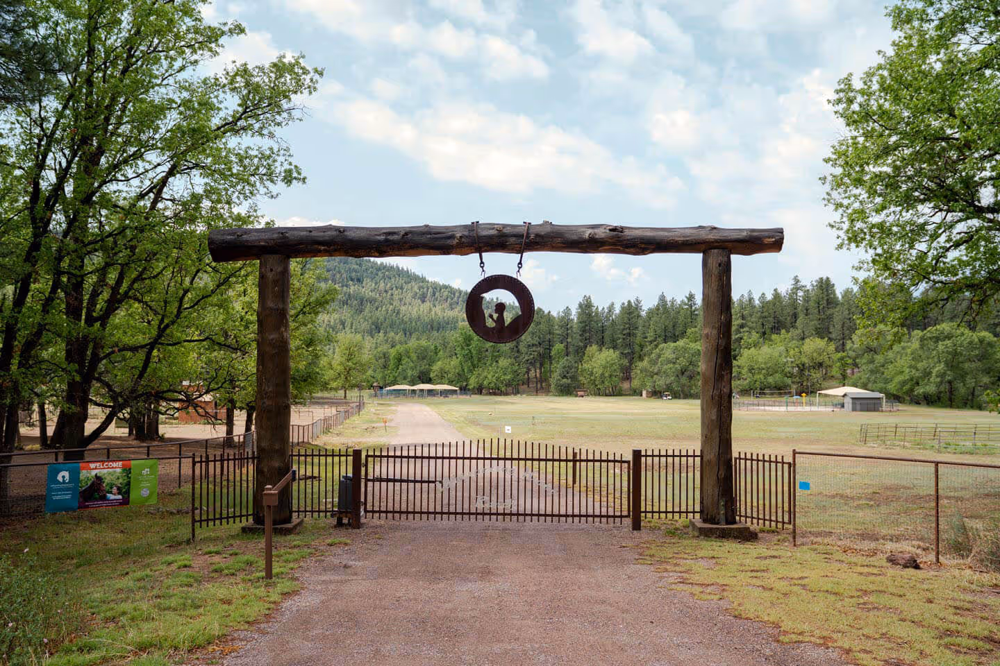 Entrance gate of Whispering Hope Ranch, made of metal bars and wooden posts with a circular hanging sign, leading to a fenced grassy area and forested hills in the background.