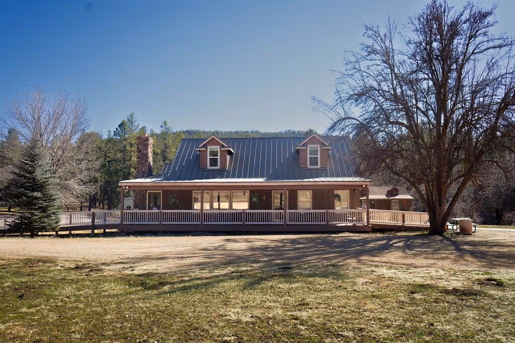 Front view of the camp's main house with a metal roof, dormer windows, wide porch, surrounded by trees and grassy yard under clear sky.
