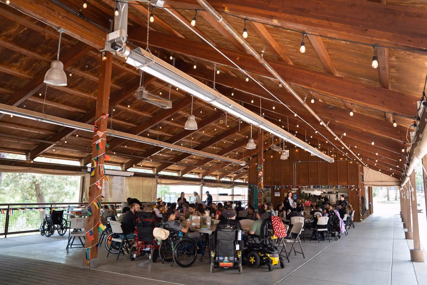 Group of people, including several in wheelchairs, seated around tables inside a spacious wooden pavilion decorated with string lights and small international flags.
