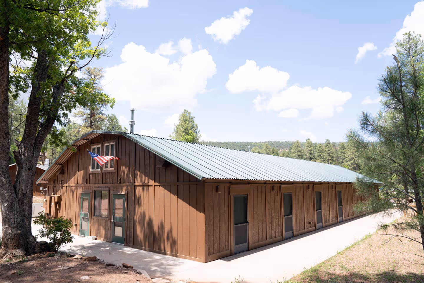 Long brown wooden bunkhouse with green doors and a metal roof, surrounded by trees under a partly cloudy sky.