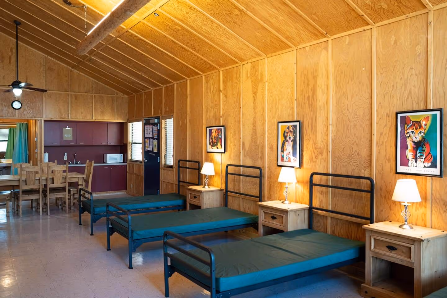 Interior of a wooden cabin room with three single metal-frame beds, each with a nightstand and lamp, colorful animal paintings on the wall, and a kitchenette with a dining table and chairs.