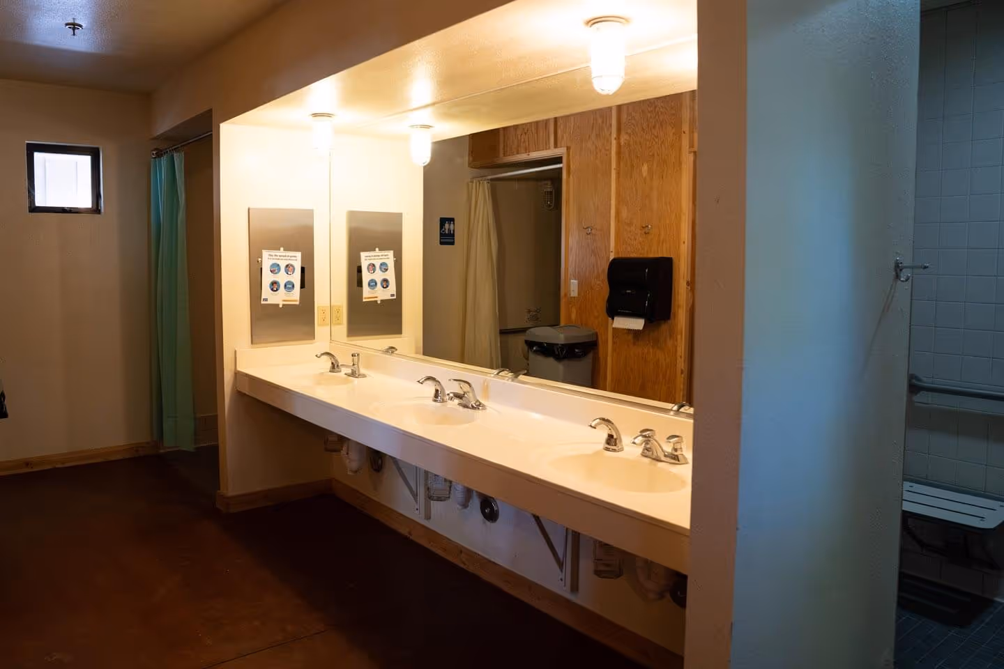 Cabins bathroom with triple sinks, large mirror, paper towel dispenser, and a shower area with a blue curtain and bench.