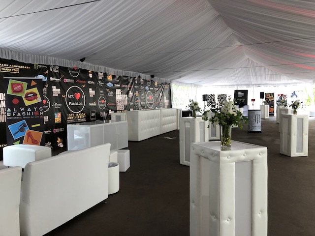 Interior of a large white event tent with tufted white furniture, small white cocktail tables with flower vases, and a black backdrop with fashion-related logos.