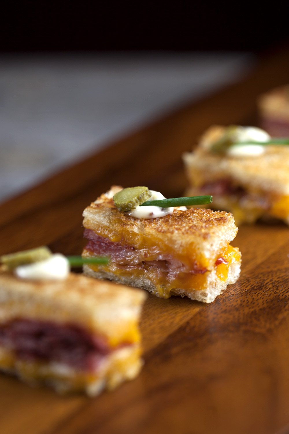 Close-up of small grilled cheese and pastrami sandwiches topped with a dollop of sauce, a pickle slice, and a chive on a wooden surface.