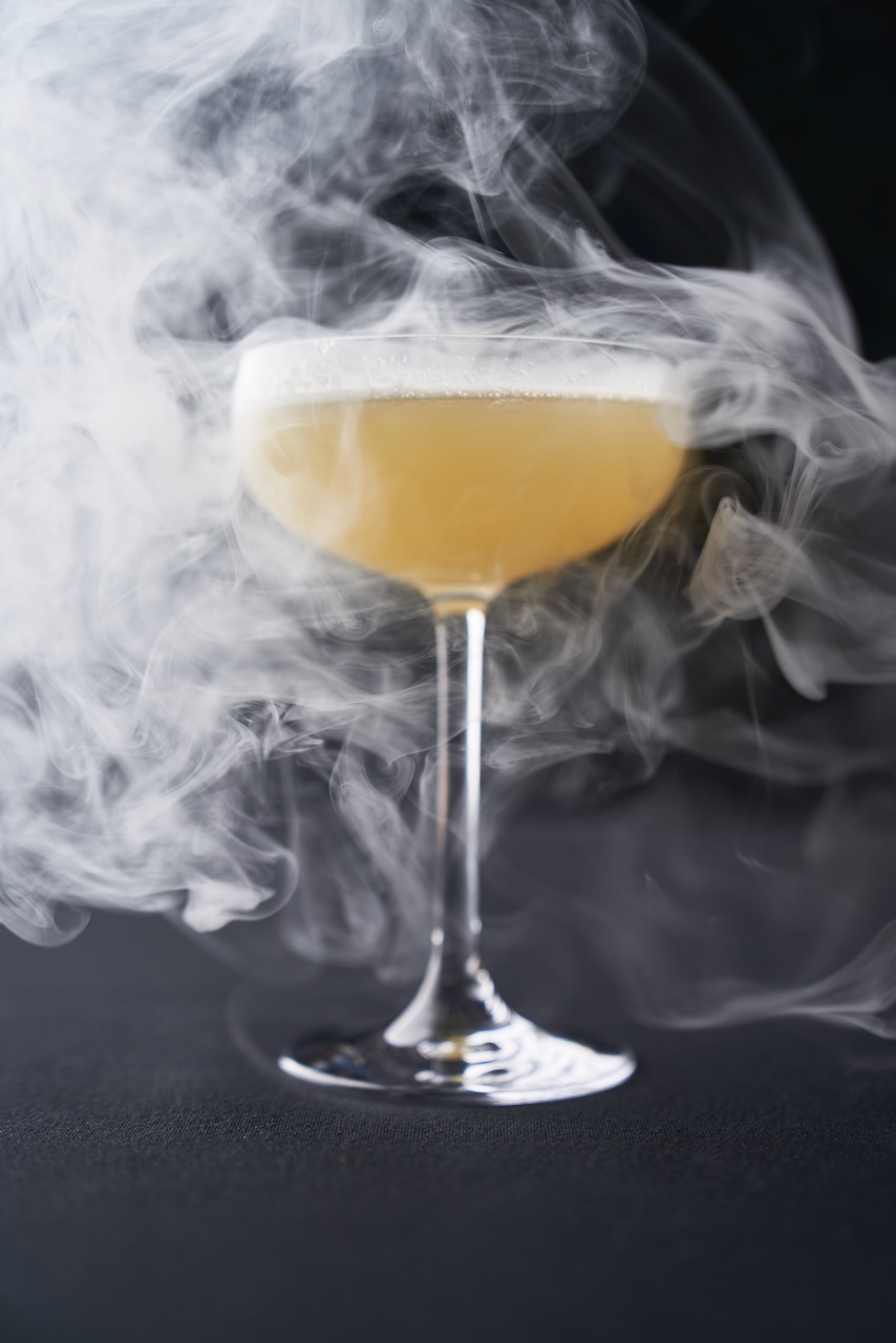 Yellow cocktail in a coupe glass surrounded by swirling white smoke on a dark surface.