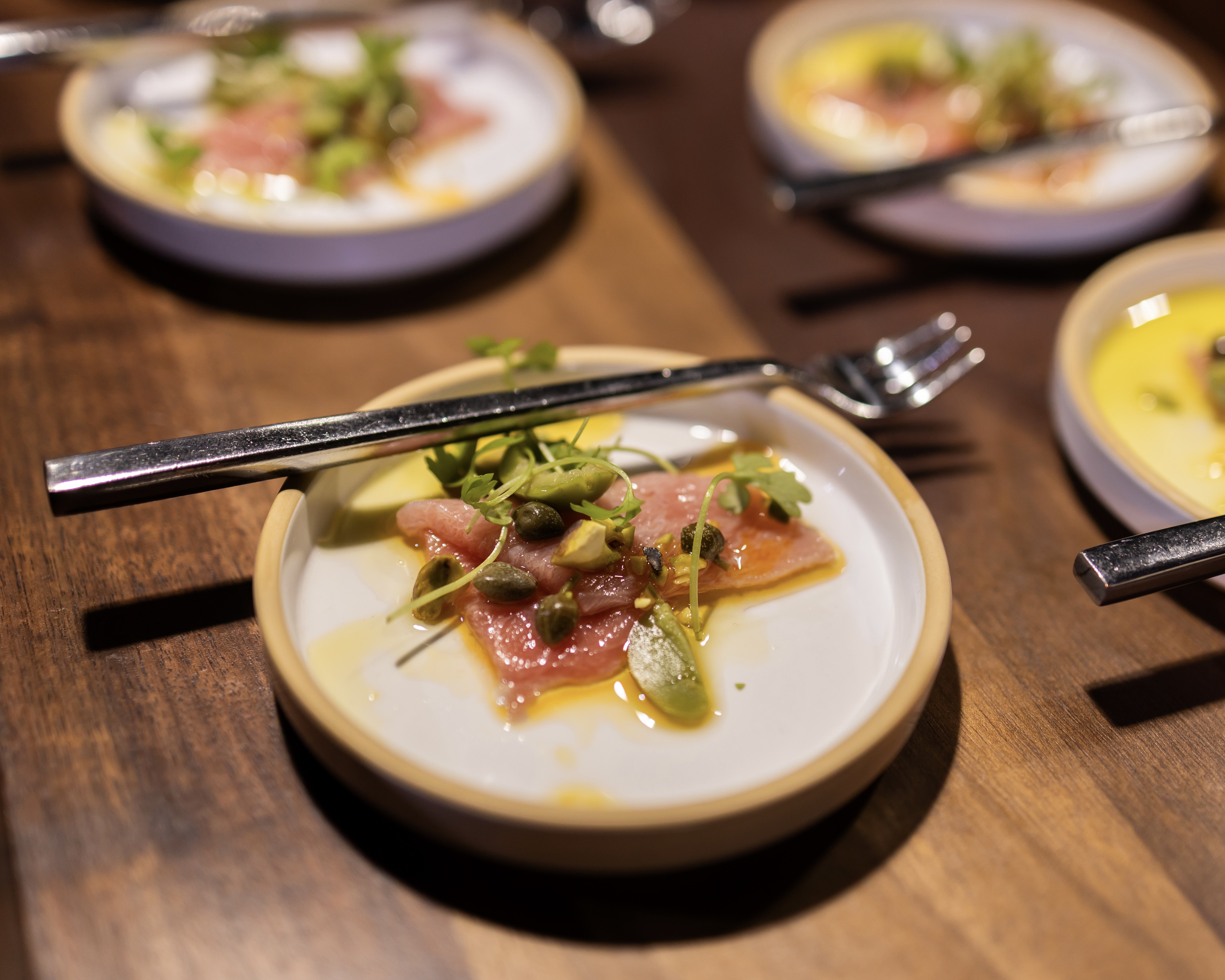 Small dish of thinly sliced raw tuna with capers, microgreens, and a drizzle of oil, served with a fork on a wooden table.