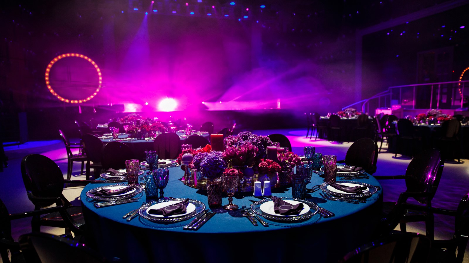 An elegantly set dinner table with pristine tableware, arranged in a sophisticated venue with a stage set up in the background, ready for a modern event to commence, showcasing the meticulous preparation involved in contemporary event planning.