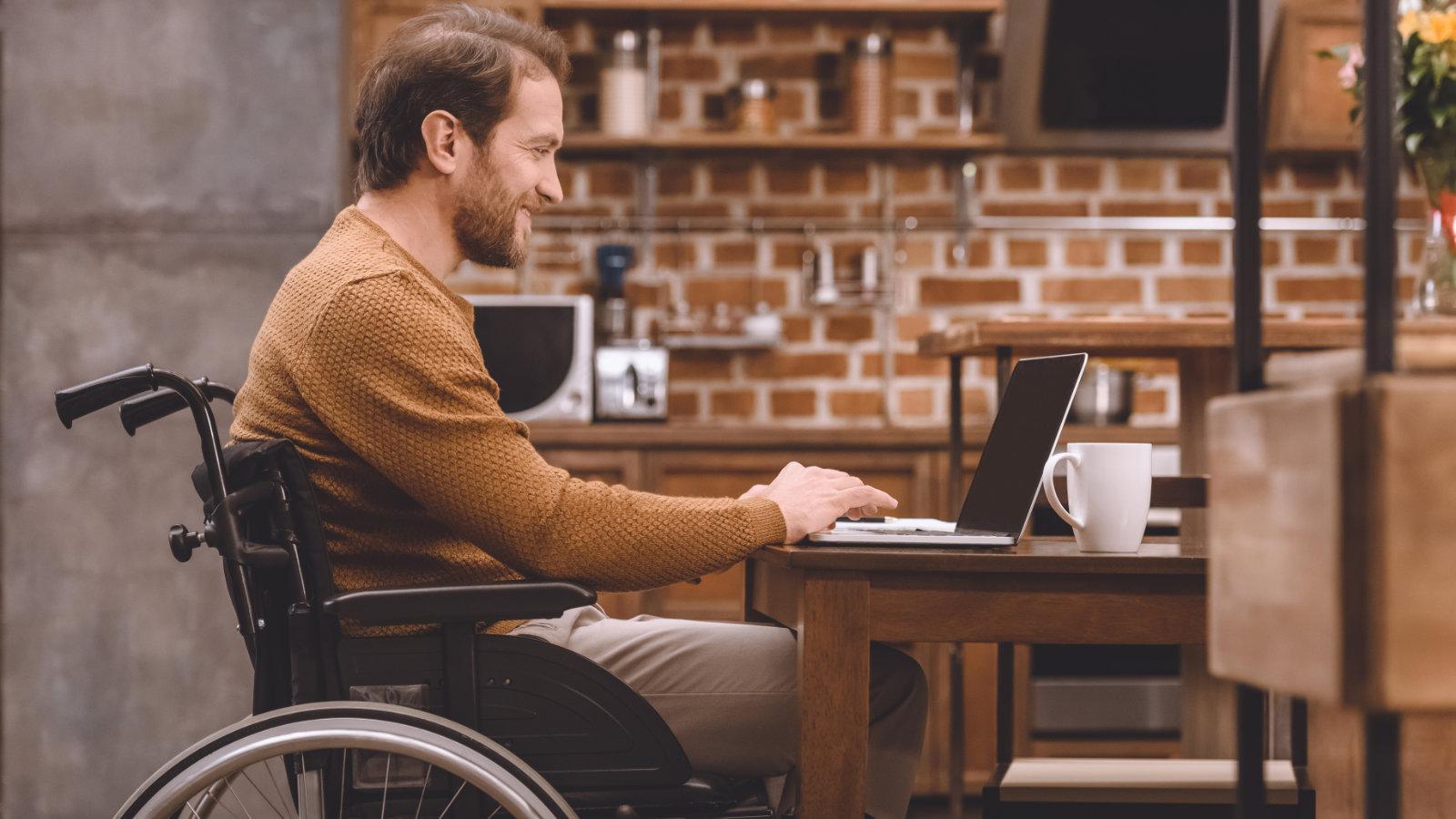 A smiling man in a wheelchair working on a laptop at a kitchen table, reflecting the inclusivity and accessibility in home design. The image evokes a sense of independence and comfort, suitable for an article discussing inclusive design principles at The