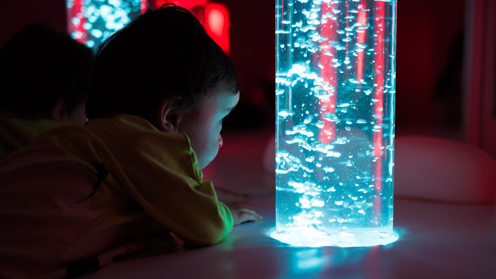 A child in a yellow shirt is mesmerized by a colorful, illuminated bubble tube, experiencing a multisensory activity in a dimly lit room.