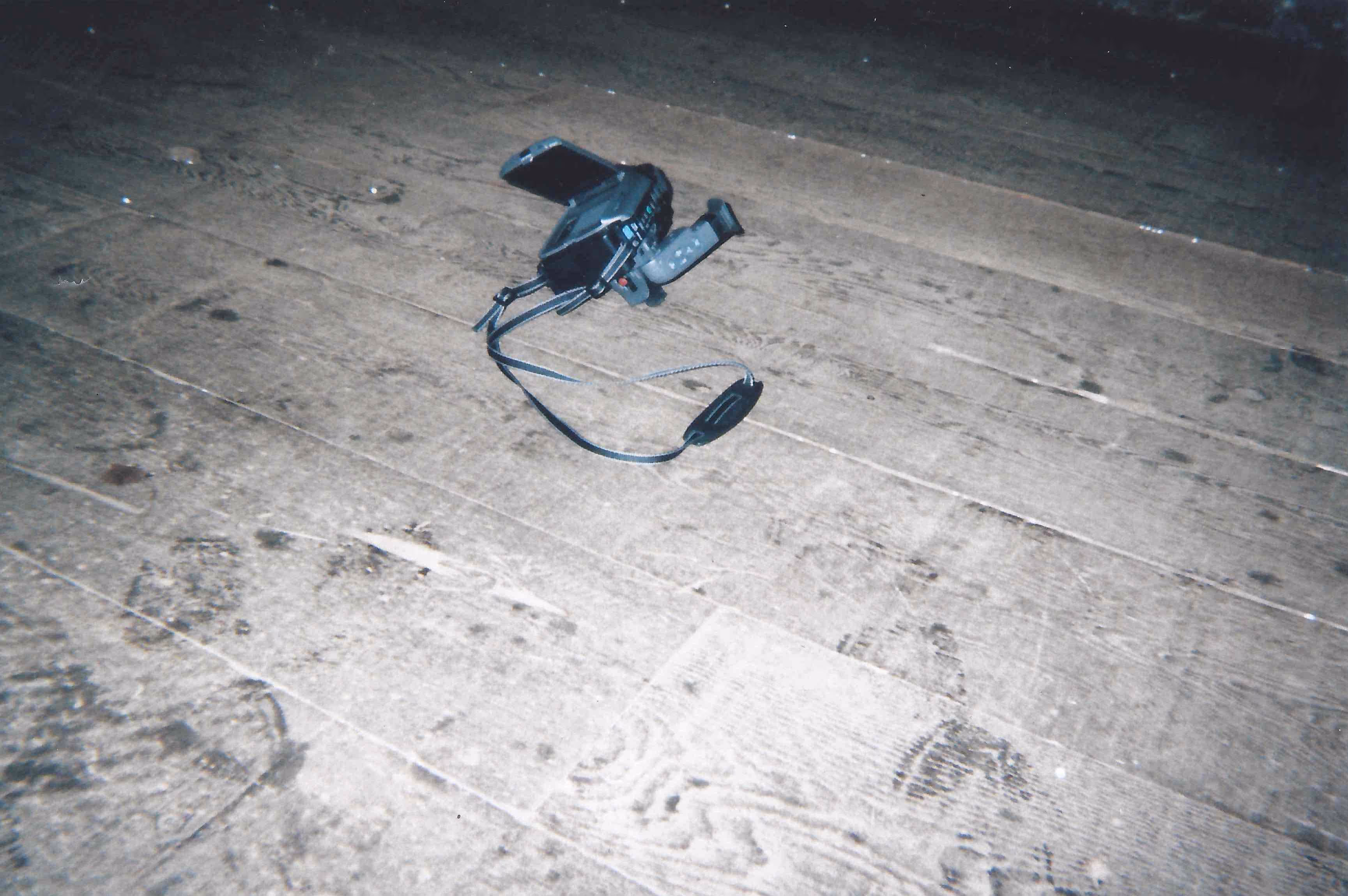 Black video camera with open screen lying on a wooden floor with visible footprints.