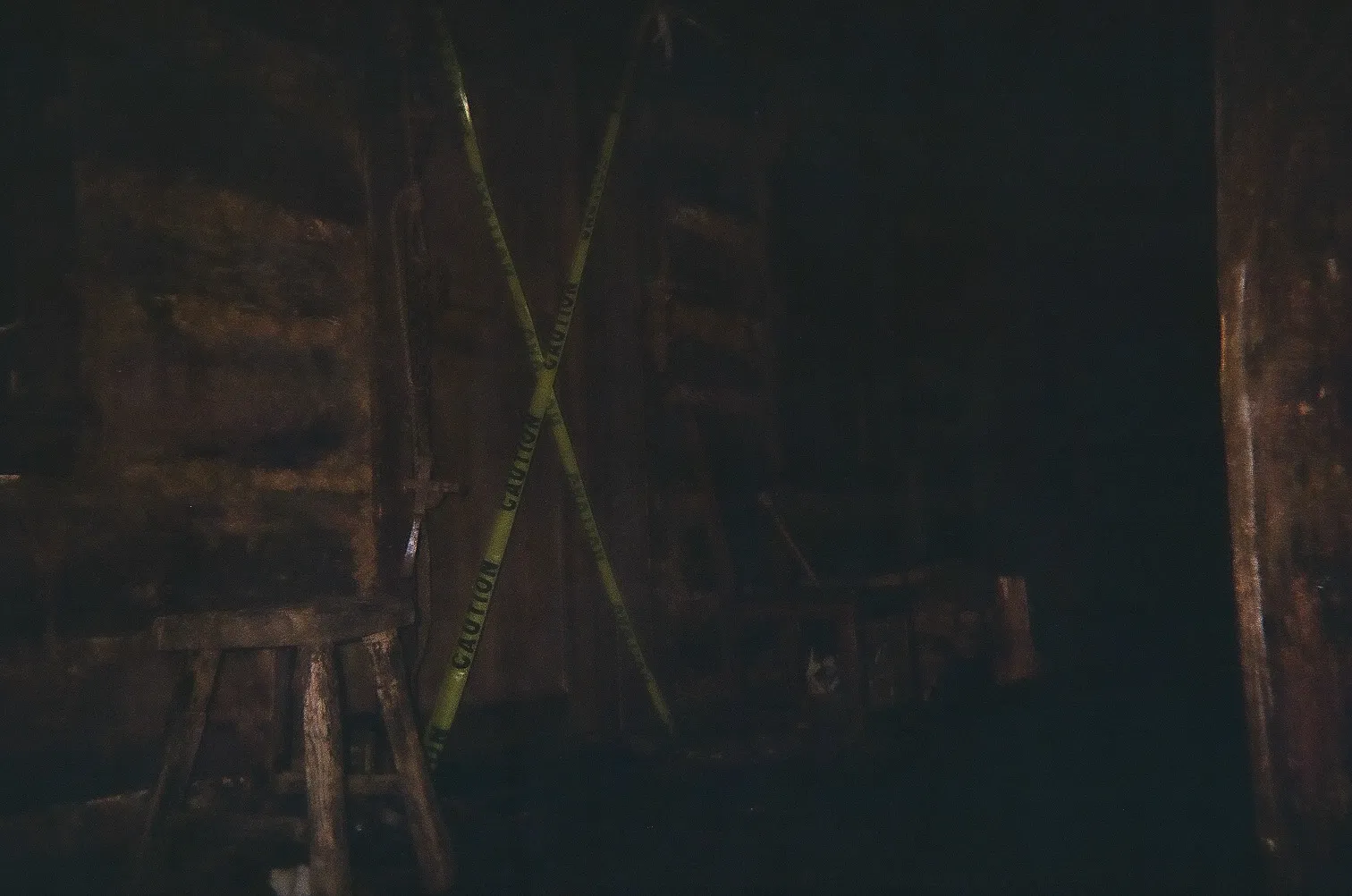 Dimly lit room with crossed yellow caution tape blocking a wooden door and rustic wooden stools nearby.