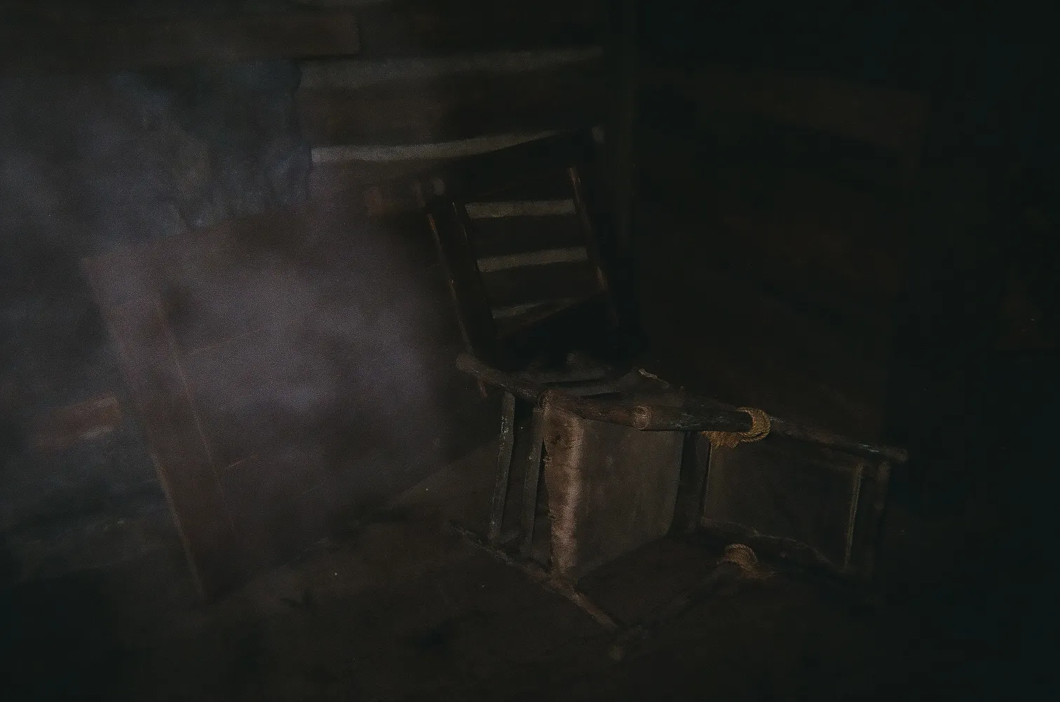 A dimly lit scene of an overturned wooden chair on a dark dirt floor inside a rustic wooden cabin.