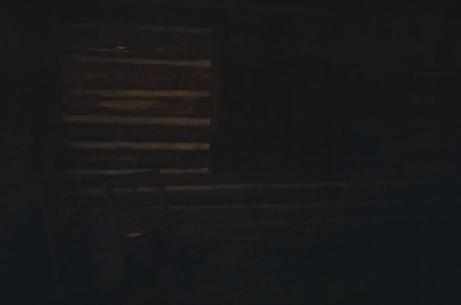 Dimly lit interior of a rustic log cabin with a wooden chair visible in the shadows.