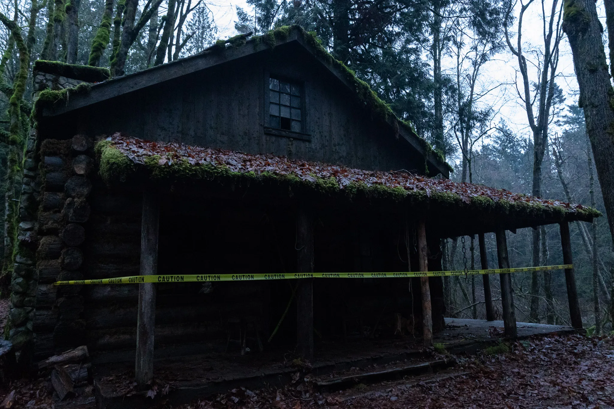 Dark wooden cabin in a forest with moss-covered roof and caution tape around the porch.
