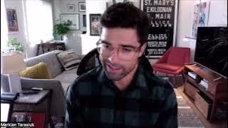 Man with glasses and dark hair wearing a dark plaid shirt sitting in a living room with a couch, red chair, and TV behind him.