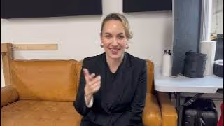 Smiling woman wearing a black blazer seated on a brown leather couch, gesturing with her right hand.