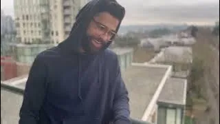 Man wearing glasses and a dark hoodie smiling on a rooftop with city buildings in the background.