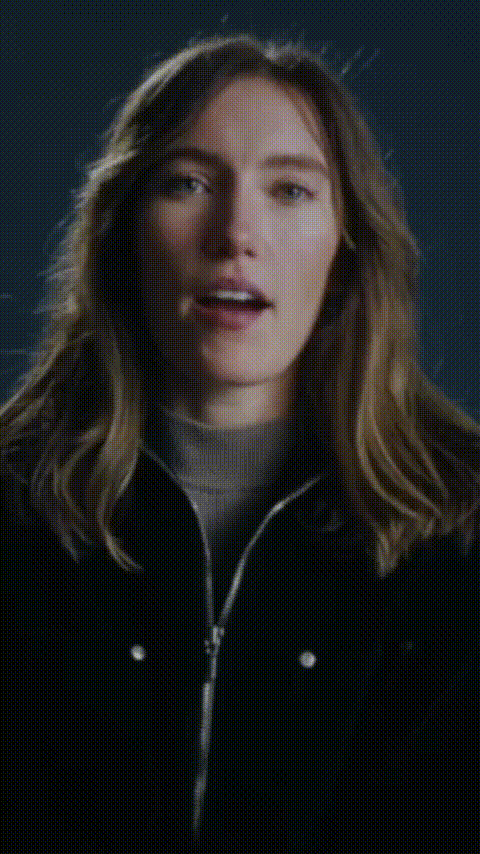 Woman with long wavy hair wearing a black jacket and gray turtleneck speaking against a dark blue background.