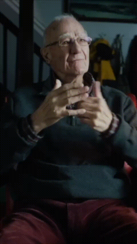 Older man wearing glasses and a dark sweater sitting and gesturing with his hands while talking indoors.