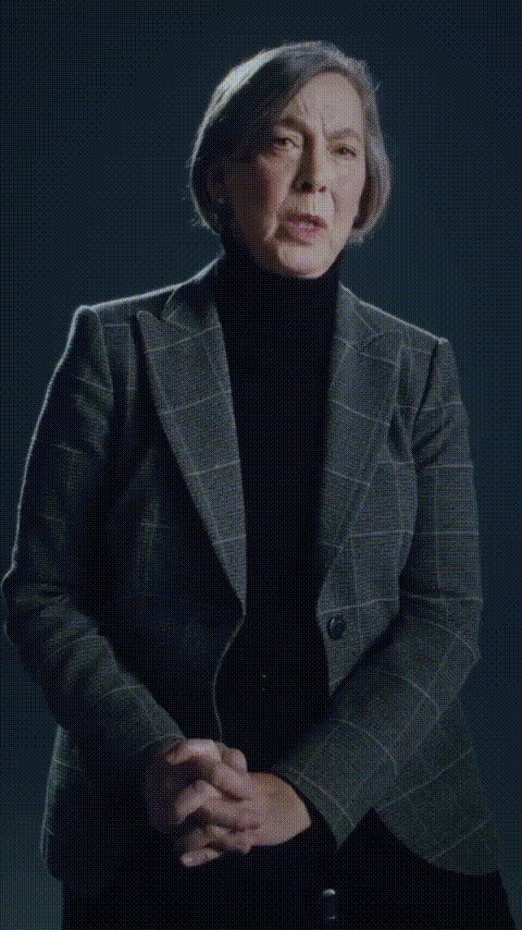 An older woman with short gray hair, wearing a checked blazer over a black turtleneck, speaking with a serious expression against a dark background.