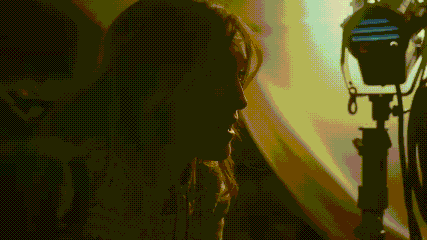 Side profile of a woman with wavy hair illuminated by soft studio lighting and surrounded by film equipment.