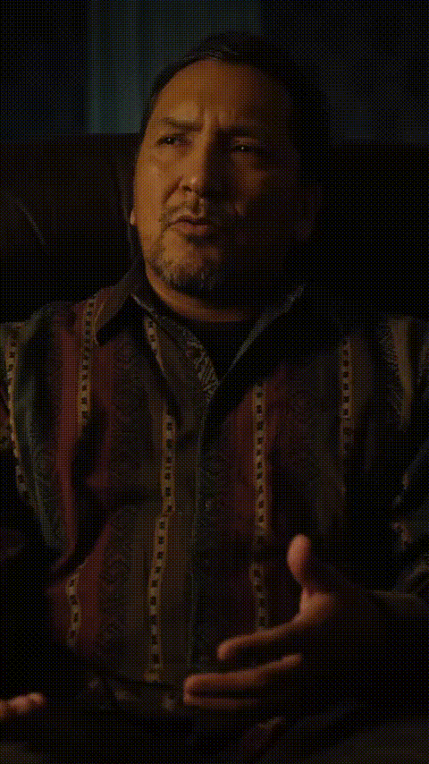 Man with short dark hair and goatee speaking and gesturing with hands in a dimly lit room.