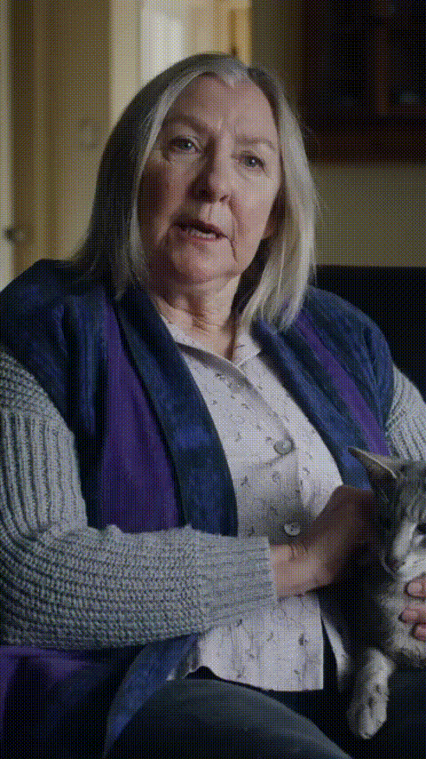 Older woman sitting on a couch petting a gray tabby cat in a cozy room.