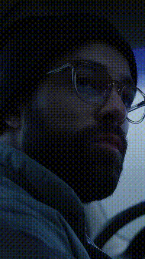 Close-up of a bearded man wearing glasses and a black beanie, looking intently to the side in a dimly lit setting.