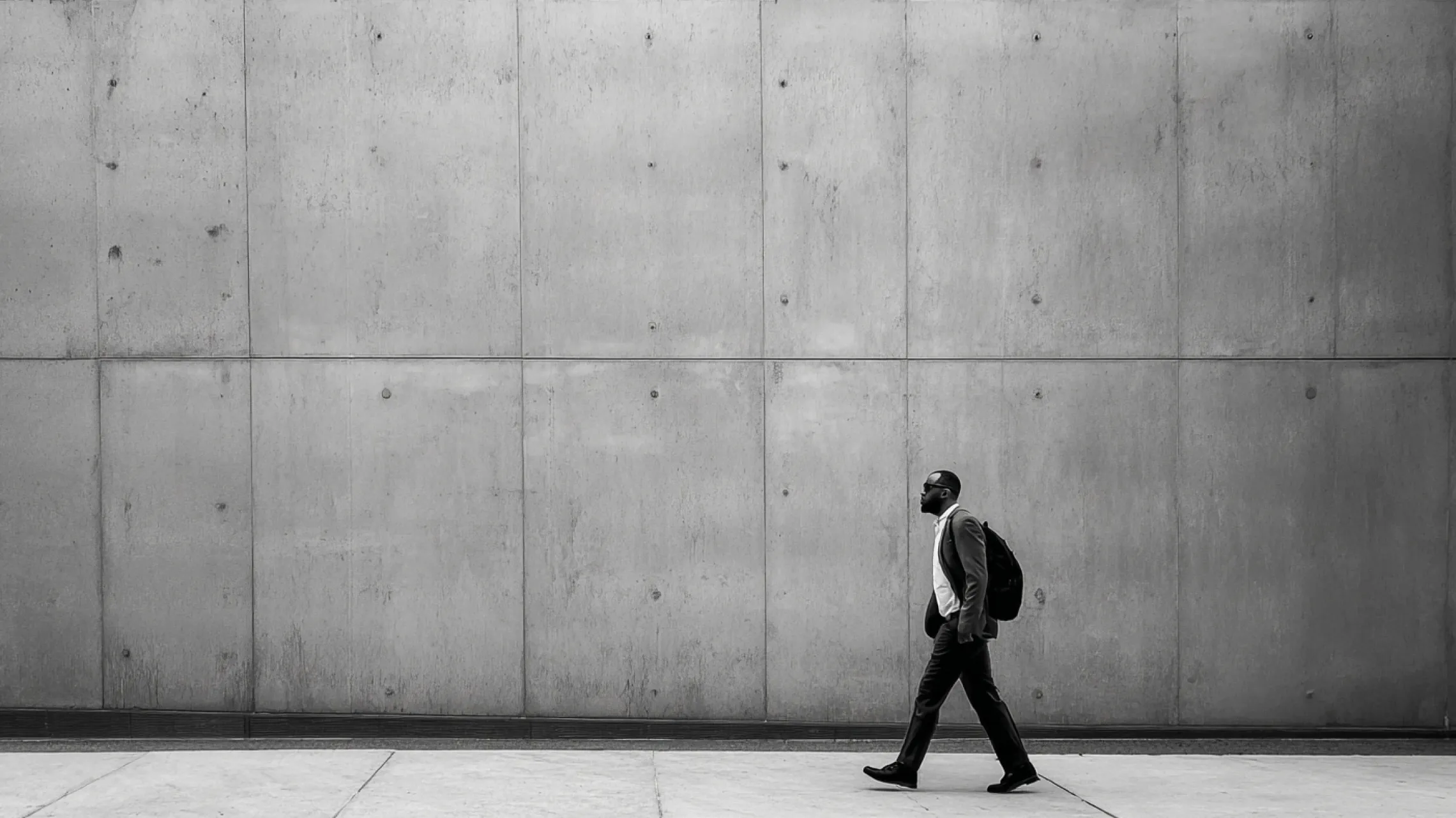 A high-contrast, black-and-white wide photograph of a man in a suit and backpack walking briskly past a massive, seamless concrete panel wall, demonstrating a human-scale element within a disciplined architectural landscape.