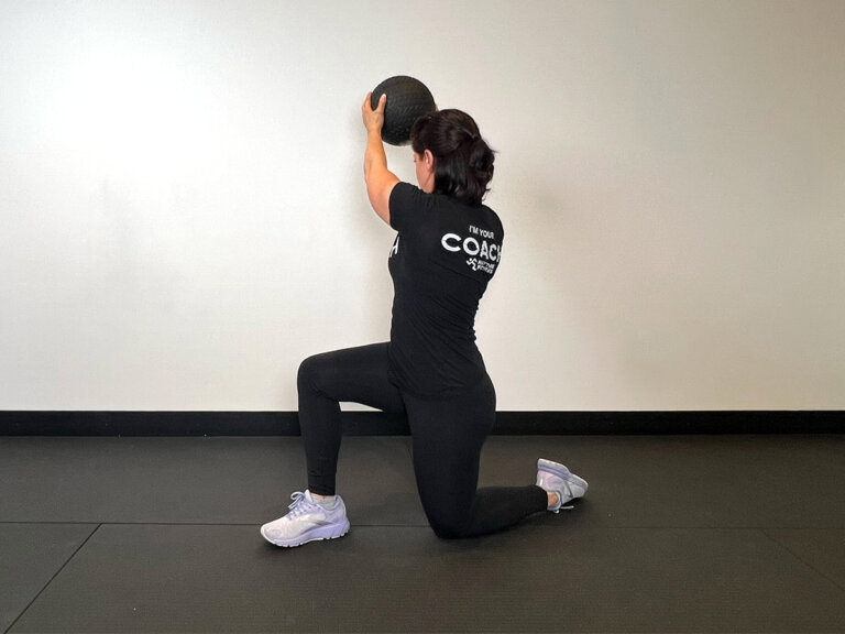 Coach in lunge position with left knee on the ground, torso twisted to the right with outstretched arms holding medicine ball up.