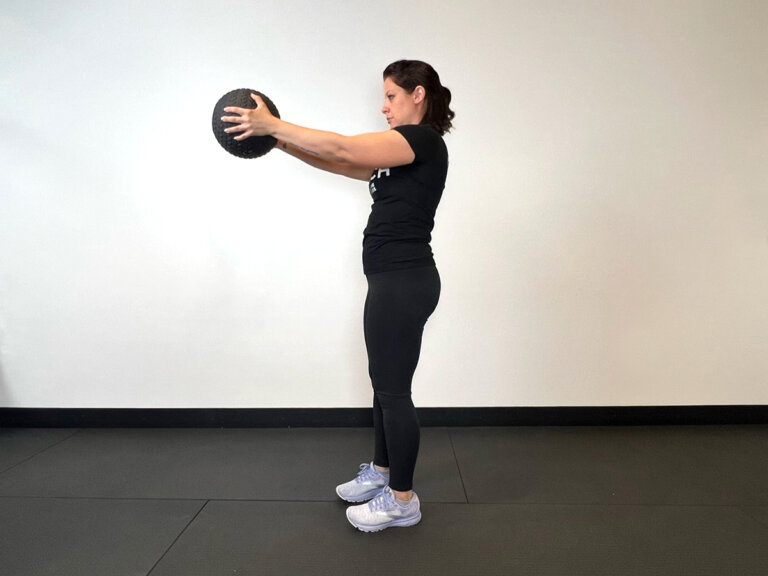 Coach stands upright while holding medicine ball straight out in front of chest.