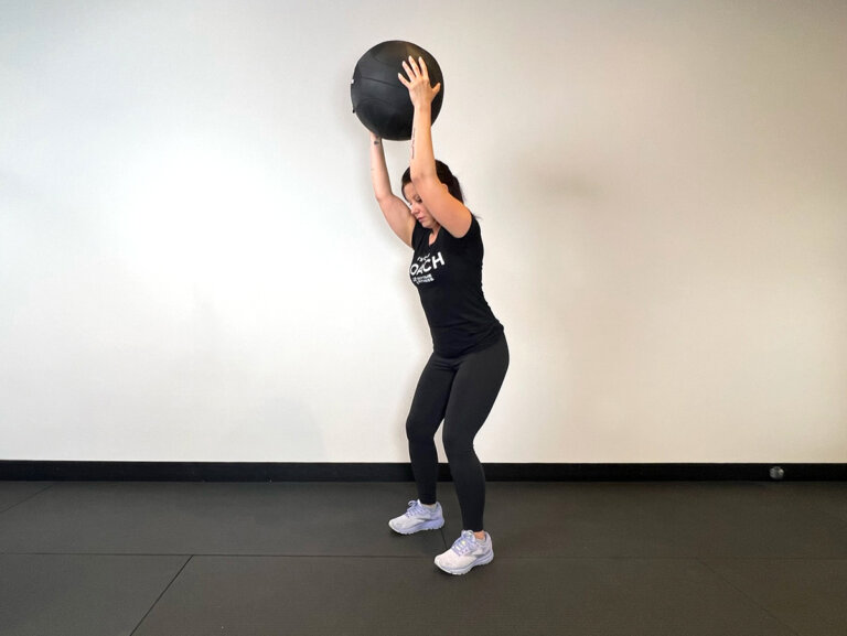 Coach stands, holding a medicine ball with both hands overhead.