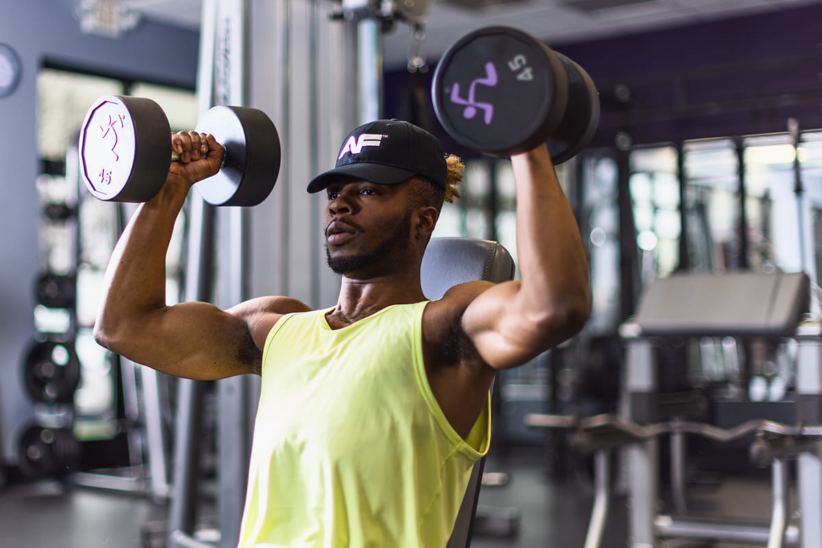 Man in a yellow tank top doing dumbbell overhead presses in an Anytime Fitness gym.