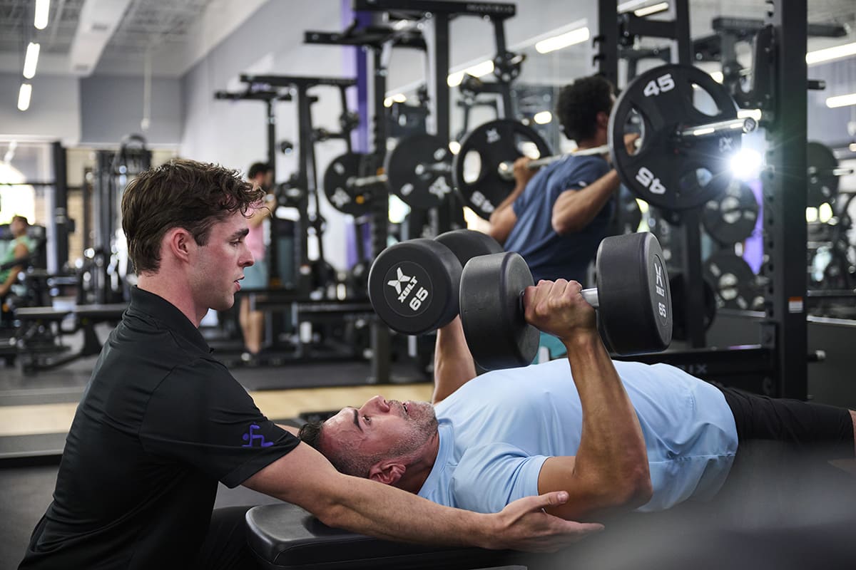 Anytime Fitness Coach helping a member do a dumbbell chest press on a bench.