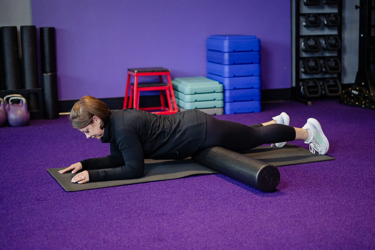 Anytime Fitness Coach demonstrating hip mobility exercises, including SMR hip stretching on a foam roller, in a gym setting.