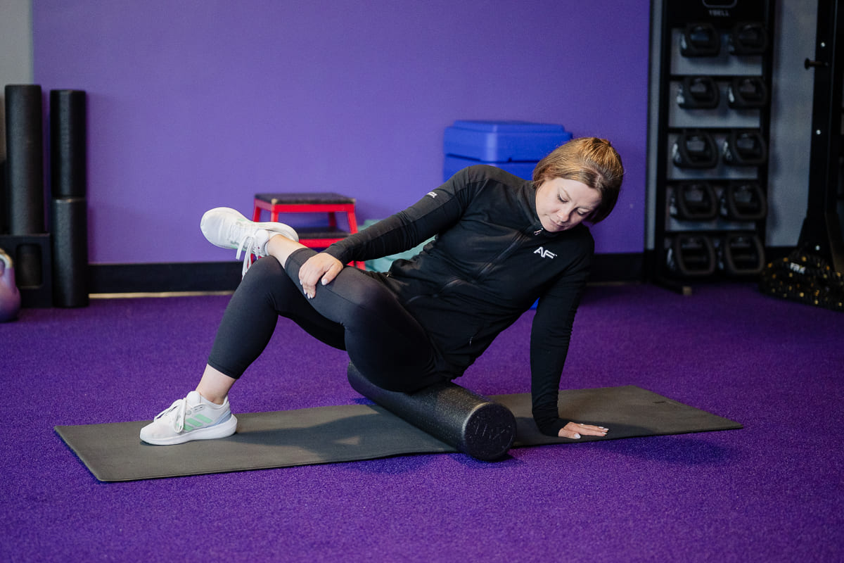 Anytime Fitness Coach demonstrating hip mobility exercises, including an SMR hip abductor stretch, in a gym setting.