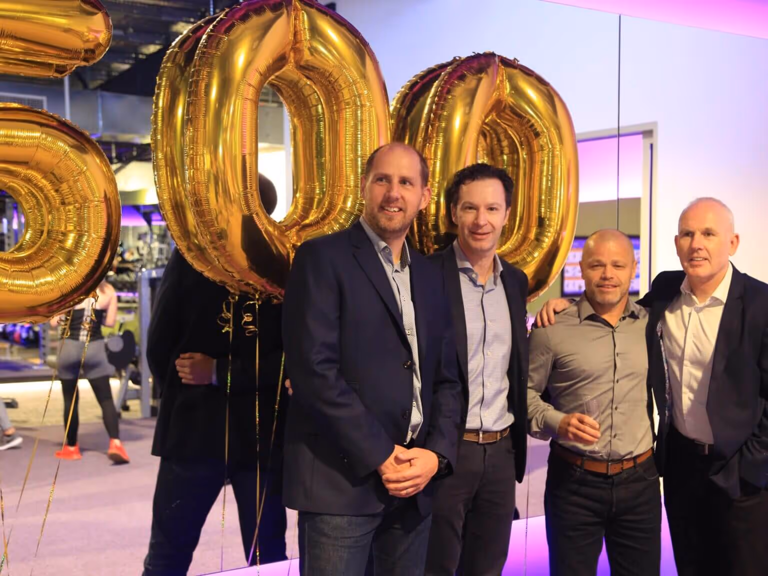 Group of people celebrating the opening of Anytime Fitness’s 500th store, holding balloons and smiling in front of gym equipment.