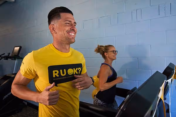 Man wearing a yellow “R U OK?” shirt smiling while running on a treadmill beside a woman exercising at the gym.