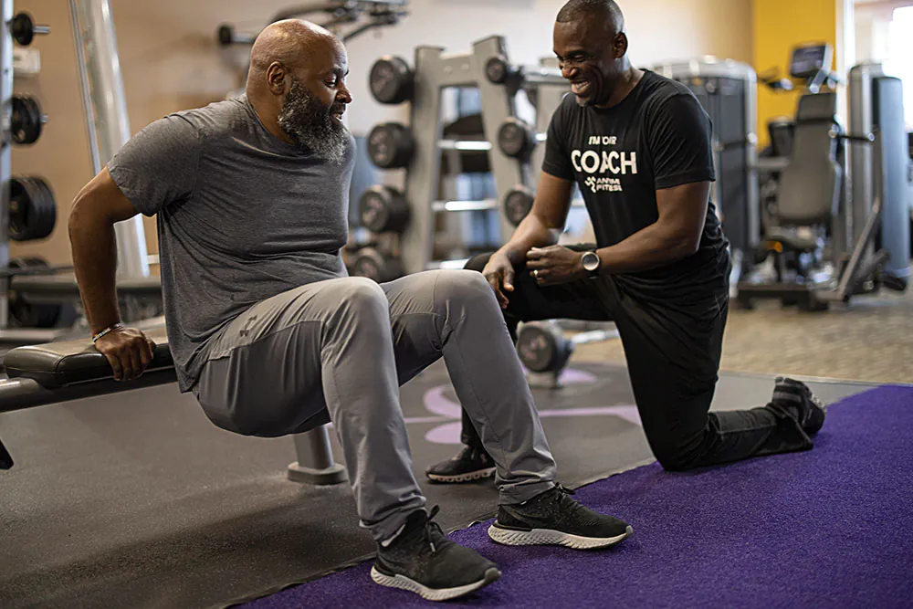 A man performs dips on a bench while a fitness coach kneels beside him, offering guidance in a gym setting filled with equipment.